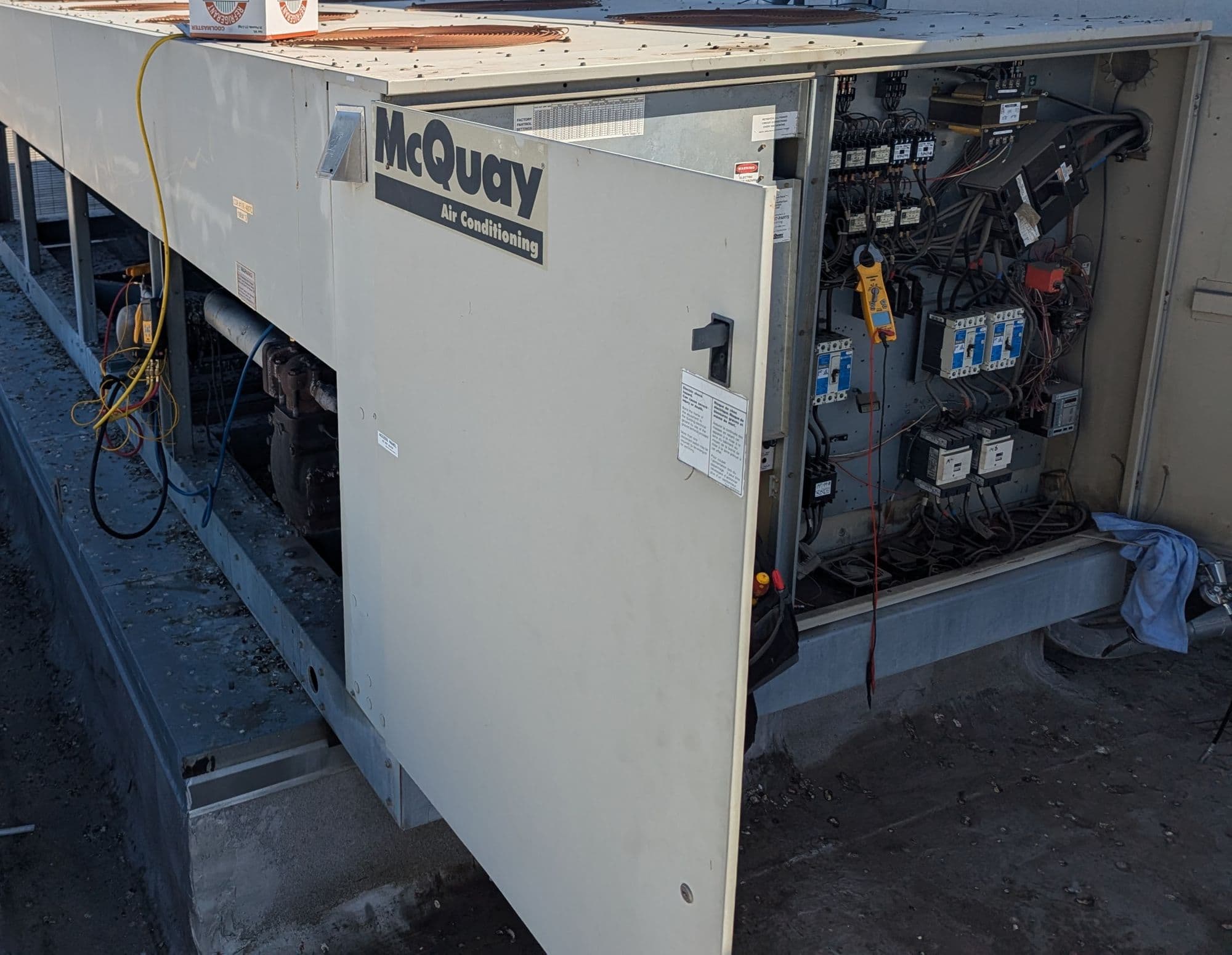 Large air-cooled condensing unit with its electrical panel open and a clamp meter on the wiring