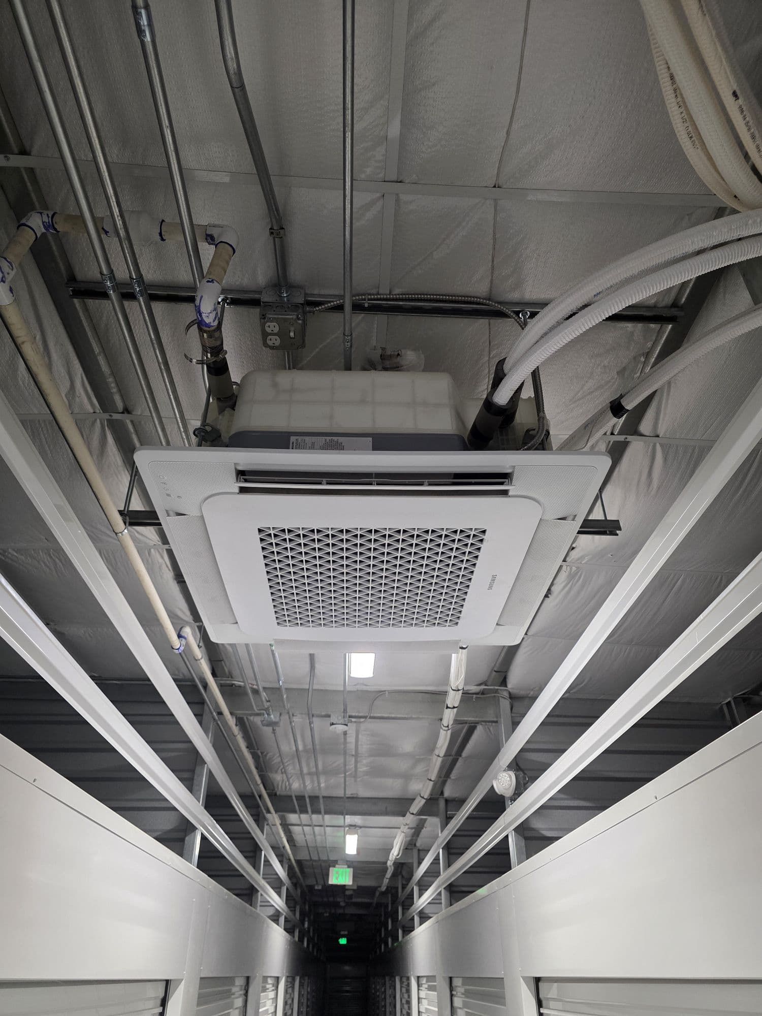 Ceiling cassette indoor unit in a commercial hallway, with refrigerant lines and conduit overhead